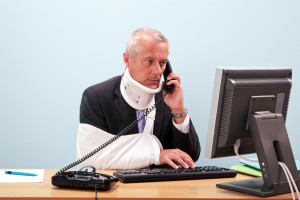 11211833 - photo of a mature businessman with injuries talking on the phone whilst trying to work on his computer. good image for health and safety or accident at work related themes.
